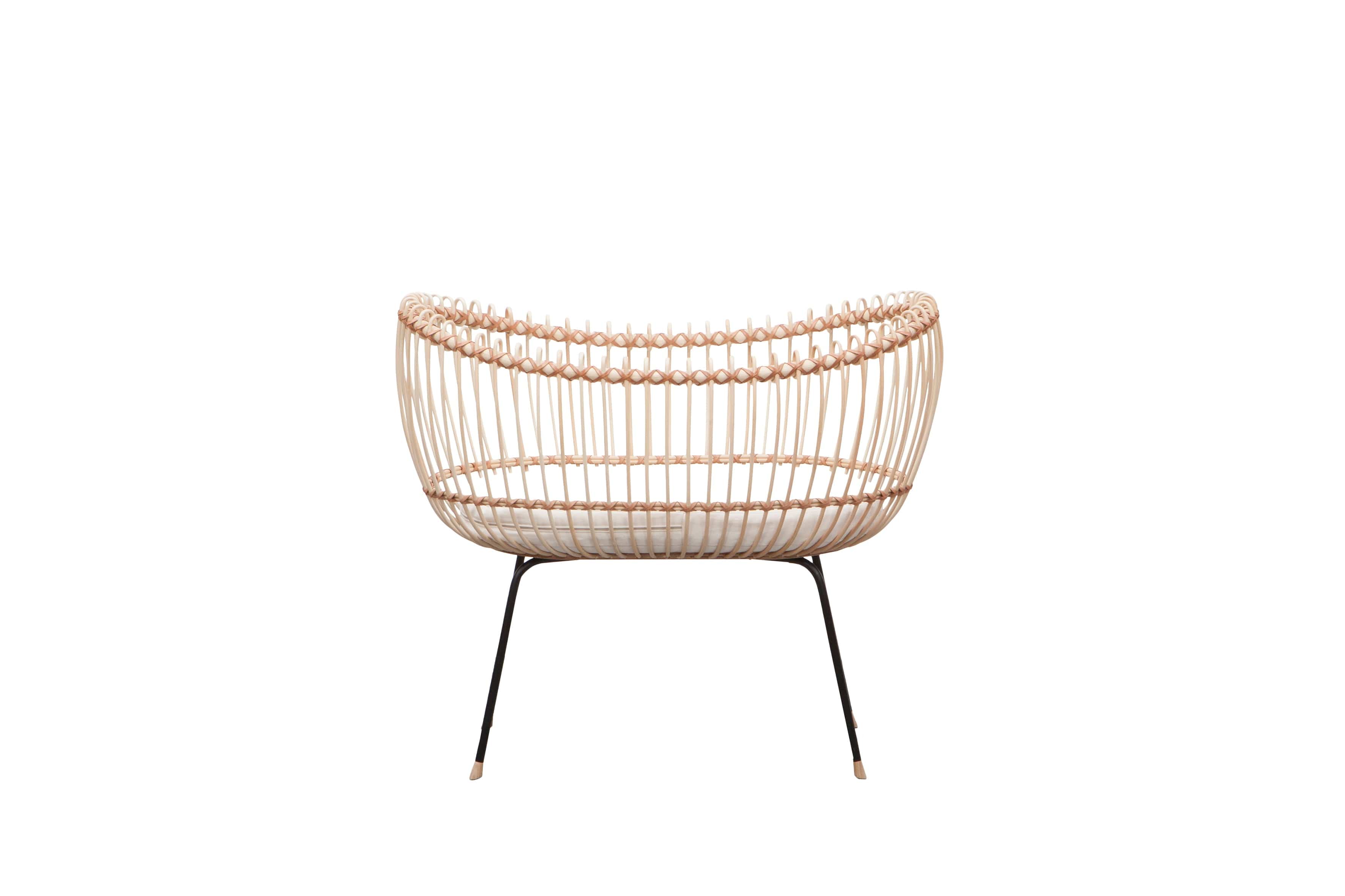 Round crib with outwards curved basket. Made of woven, light beige rattan beams, connected with light brown leather straps and standing on slim black metal legs.