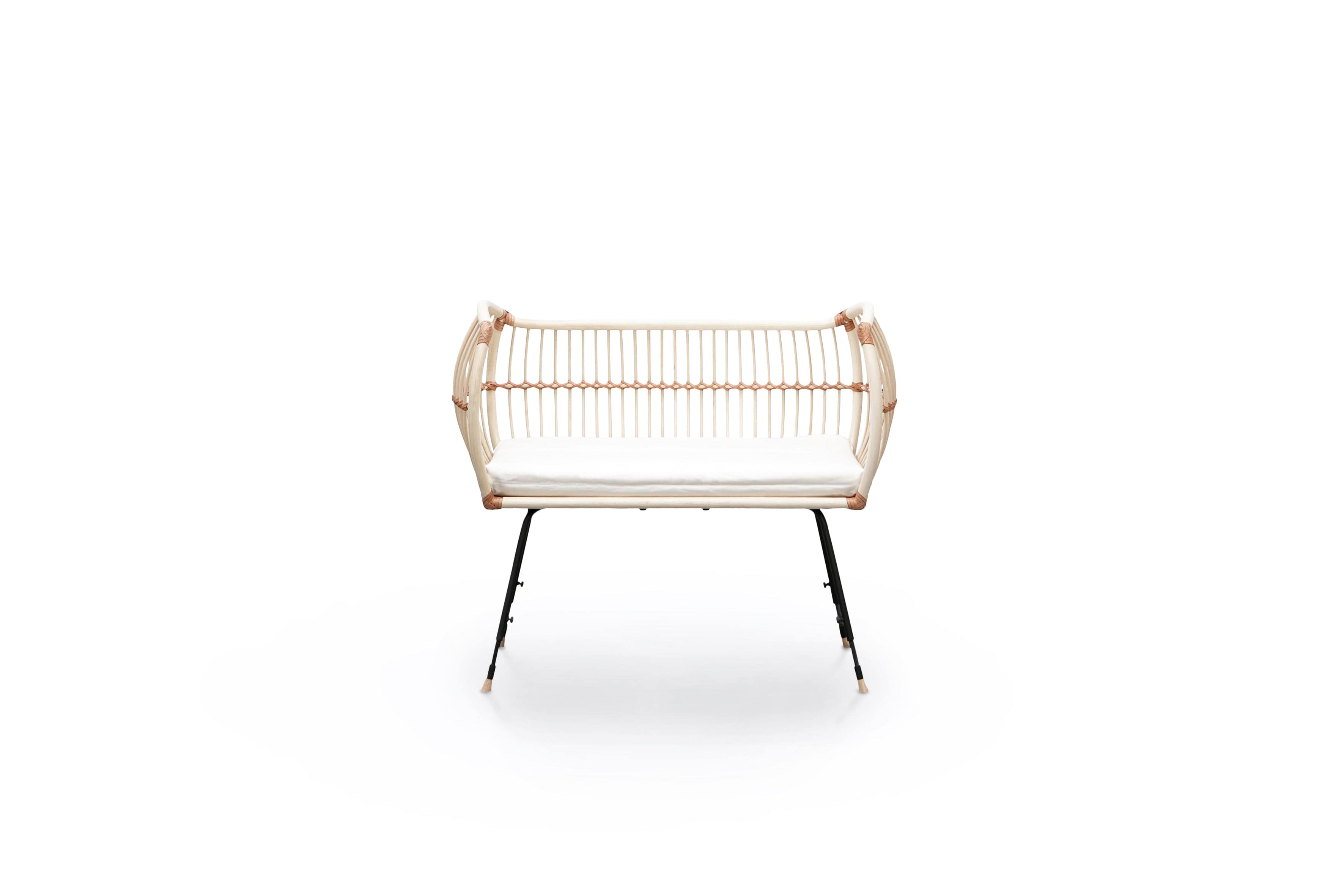 Square bed with outwards curved basket, open on one side. Made of light beige rattan beams, connected with light brown leather straps and standing on slim black metal legs. 