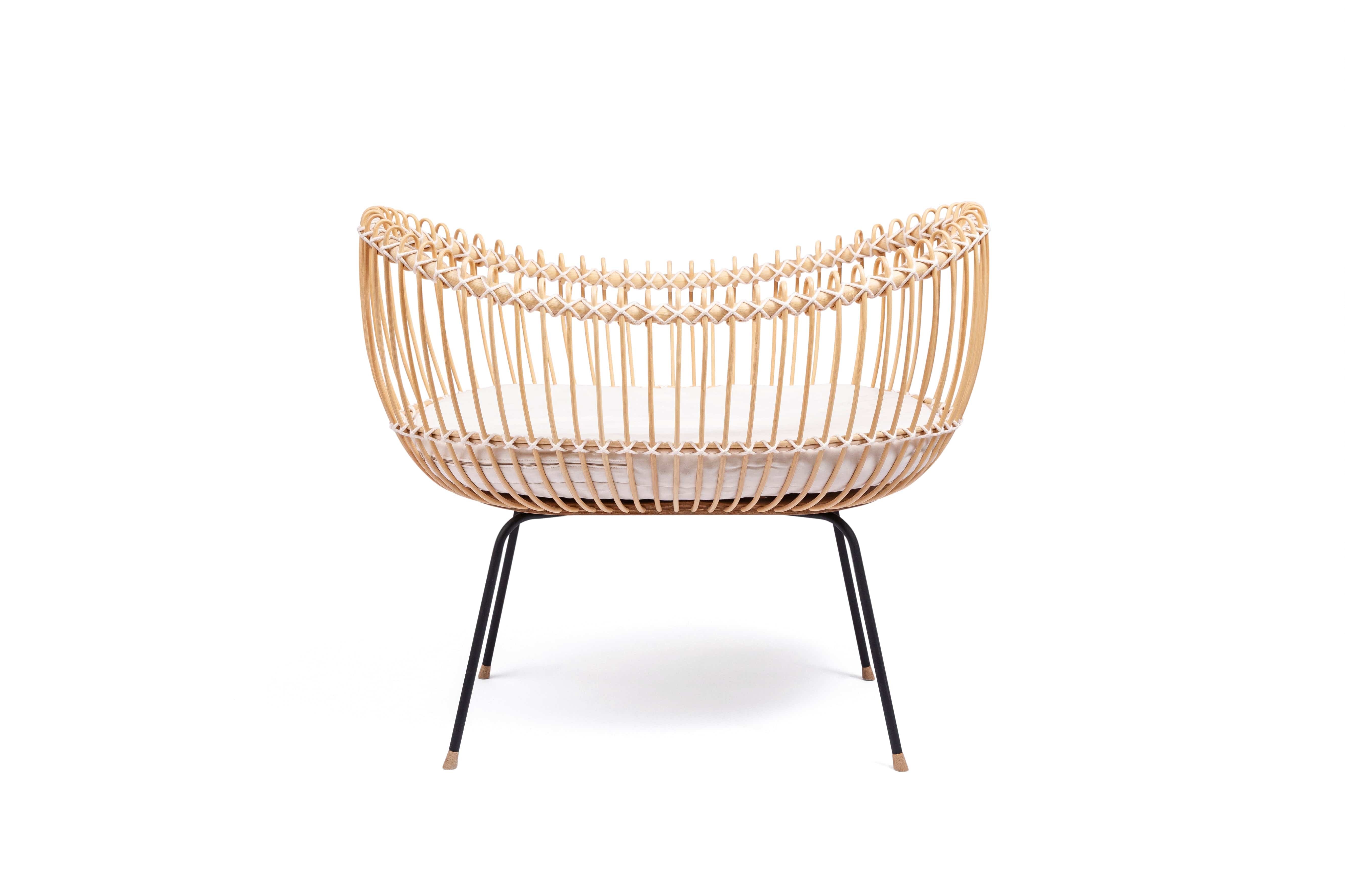 Round crib with outwards curved basket. Made of woven, light beige rattan beams, connected with off-white cotton strings and standing on slim black metal legs.