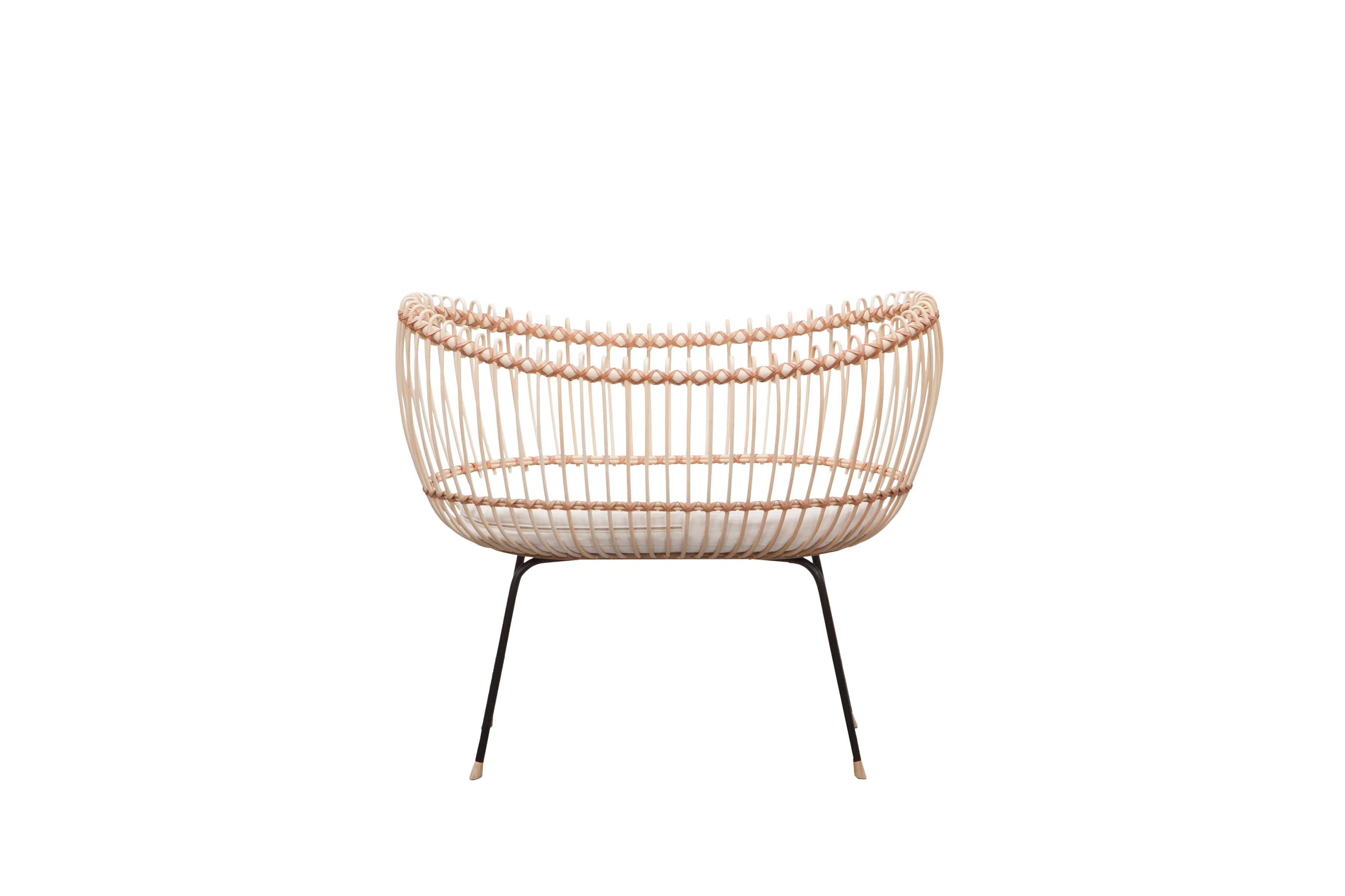 Round crib with outwards curved basket. Made of woven, light beige rattan beams, connected with light brown leather straps and standing on slim black metal legs. 