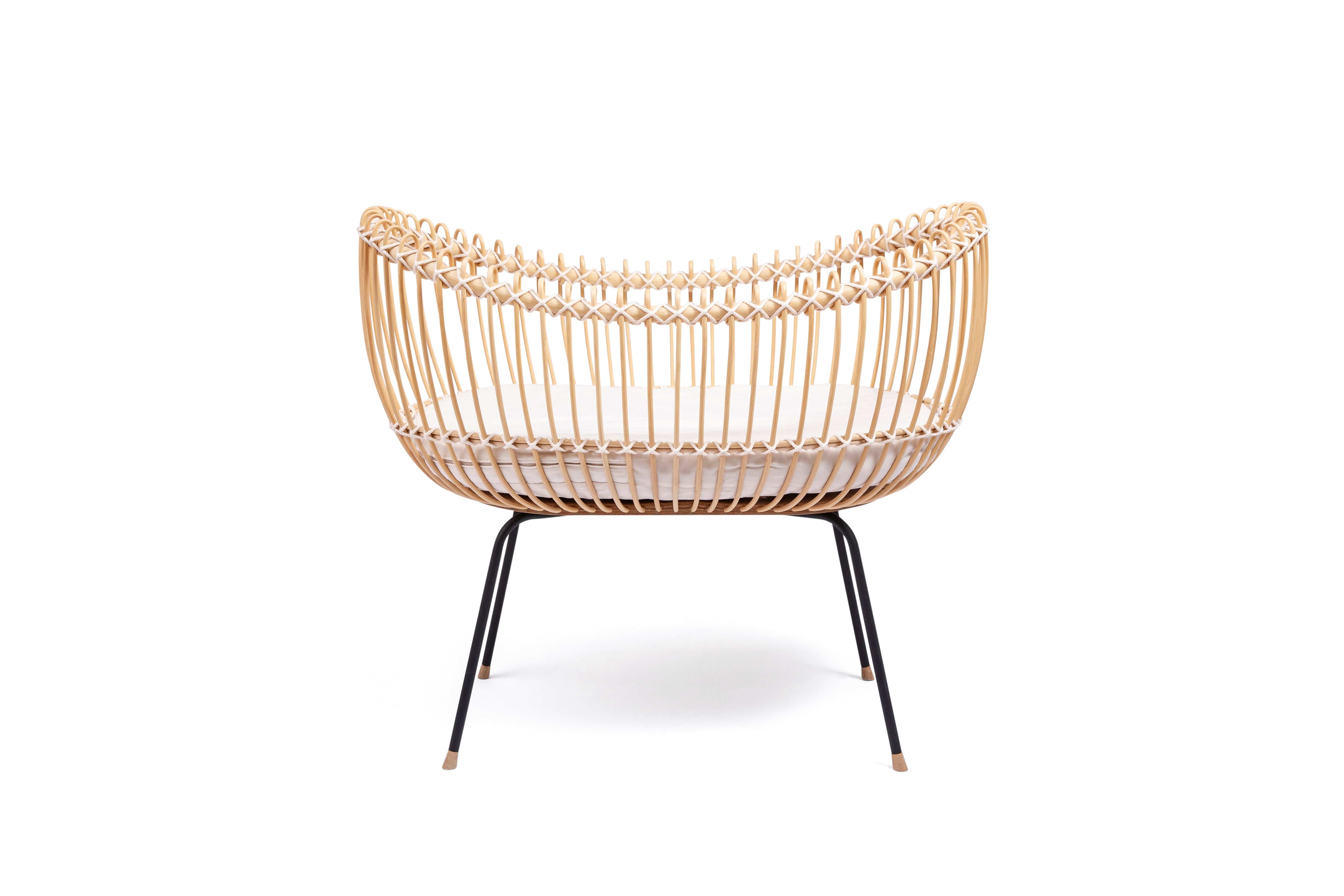Round crib with outwards curved basket. Made of woven, light beige rattan beams, connected with off-white cotton strings and standing on slim black metal legs. 
