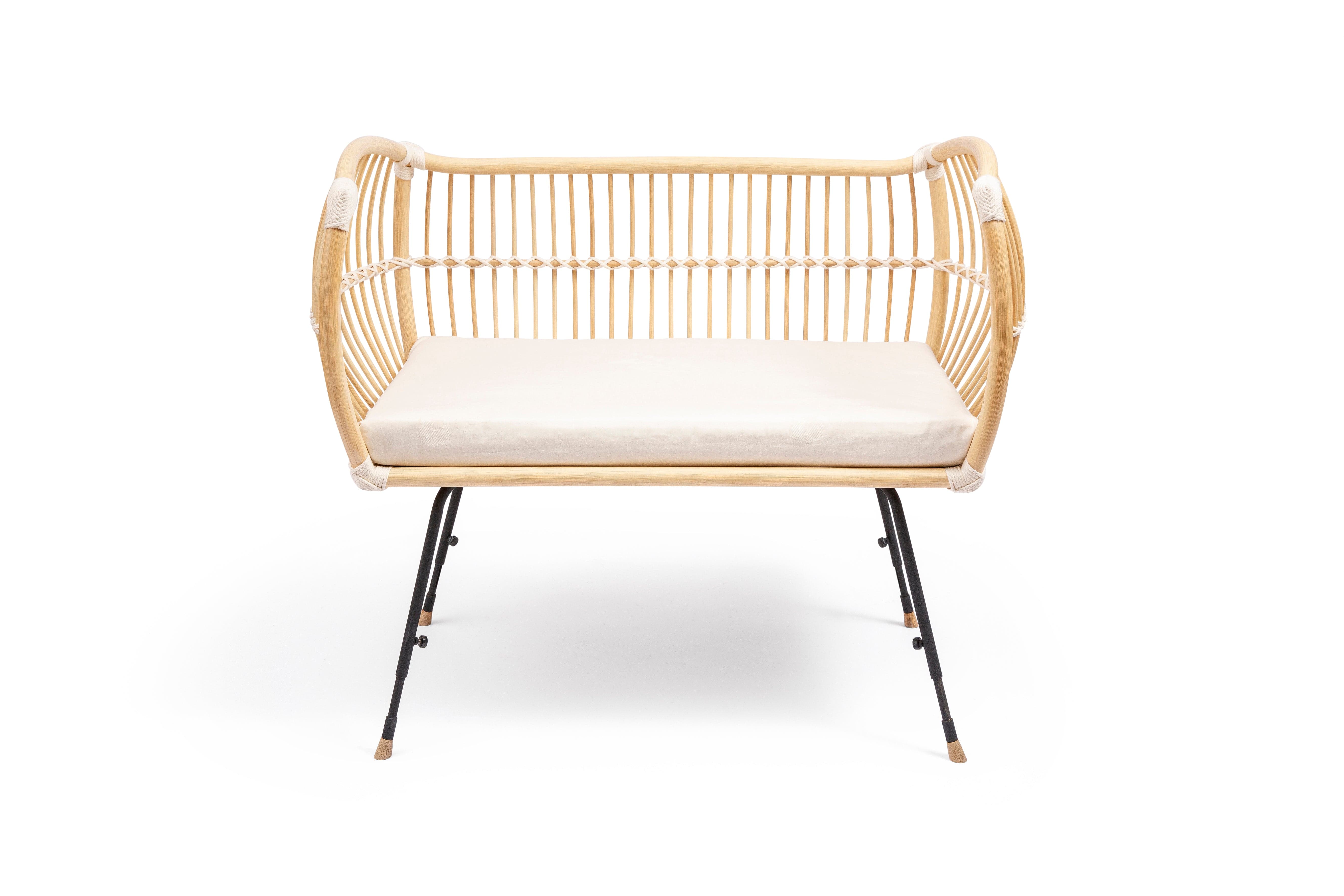 Square bed with outwards curved basket, open on one side. Made of light beige rattan beams, connected with off-white cotton strings and standing on slim black metal legs. 